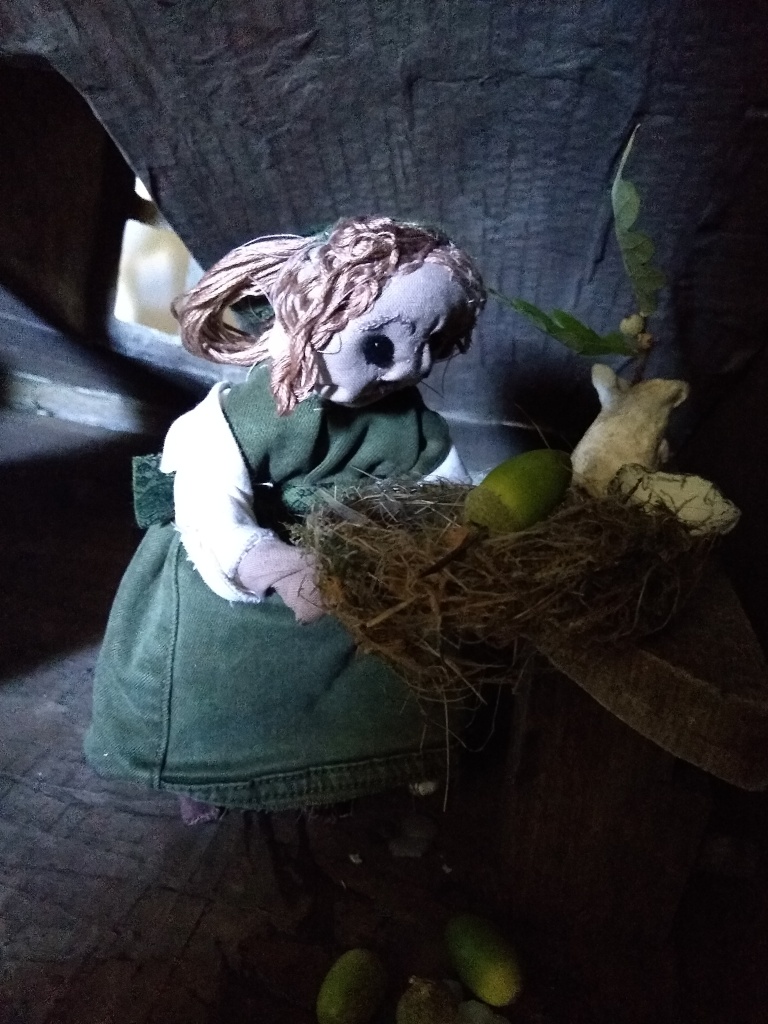 Wearing a Victorian-style green dress, a girl puppet made from cloth holds a birds nest. One green acorn rests in the nest, others have fallen onto the attic floor.